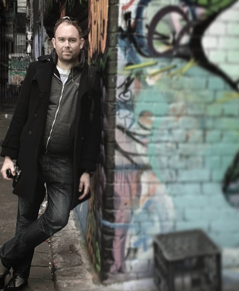 James Ian Gray leaning against a graffiti-covered brick wall in an alley, holding a camera
